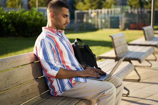Handsome middle eastern programmer using laptop computer working freelance project online at workplace. Iranian student studying sitting on bench in university campus