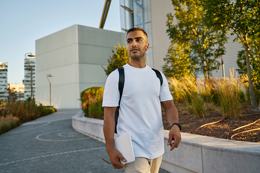 Young handsome middle eastern man wearing white t shirt walking on the street looking away. Portrait of smiling successful student with backpack in university campus
