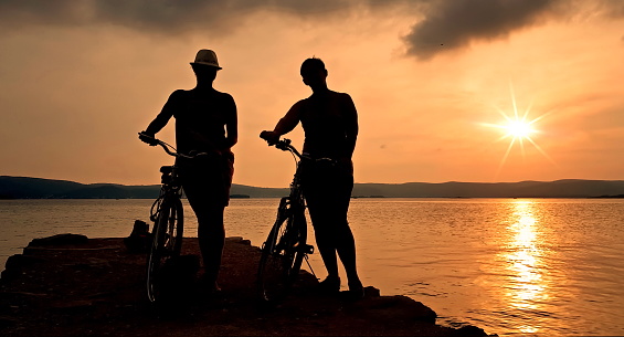Silhouette of two women with bicycles on the rock against sea sunset
