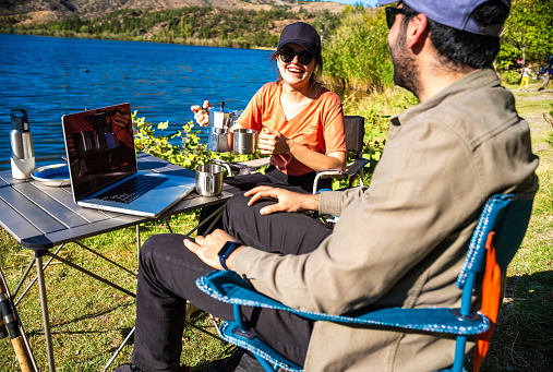 A man and a women are drinking coffee in camping . The woman is pouring coffee in her coffee cup. They are sitting beside the lake