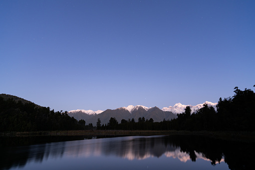 As evening g sets in, the stars appear overhead and are reflected in the mirror-like surface of spectacular Lake Matheson on New Zealand’s South Island. Beyond we see the snow capped peaks of Mt Tasman and Mt Cook and the Southern Alps.