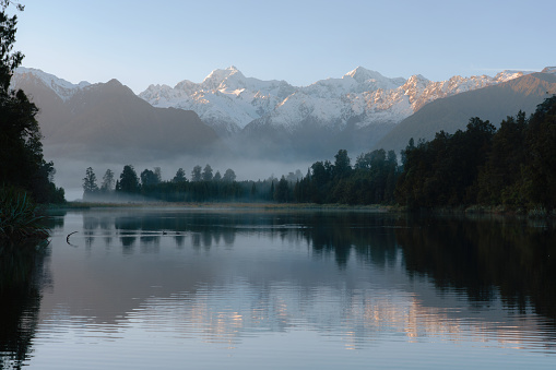 Dawn breaks over spectacular Lake Matheson in New Zealand, whilst beyond we see the snow capped peaks of the Southern Alps. A duck paddles across the mirror-like surface of the lake.