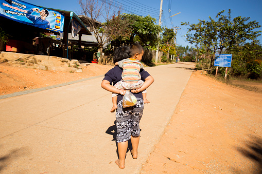 Chiang Mai, Thailand; 1st January 2023: A woman carrying a child on her back and a bag of food in a village in the mountains of Chiang Mai..