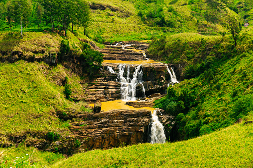 A waterfall cascading down a series of rocky steps in a lush green valley. The waterfall is the focal point of the image, with its light brown water contrasting against the darker brown rocks. The surrounding greenery, including trees, bushes, and grass, adds to the tranquility of the scene. The sky is not visible in the image, but the overall mood of the image is peaceful and serene. The photograph is taken from a distance, allowing viewers to take in the entire waterfall and surrounding landscape.