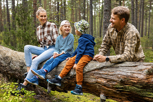 happy family with kids sitting and resting on fallen tree after walk in forest. outdoor activities, nature adventure