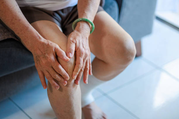 starsza azjatka masuje kolano, siedząc na sofie w salonie w domu, wyraźnie cierpiąc na ból kolana - arthritis senior adult rheumatoid arthritis sadness zdjęcia i obrazy z banku zdjęć