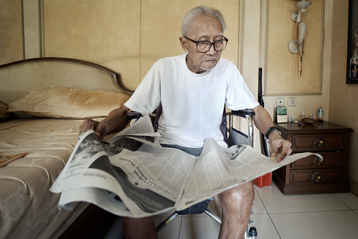 JAKARTA - Indonesia. September 15, 2023: Asian man in his 80s reading newspaper sitting on the wheelchair in the bedroom