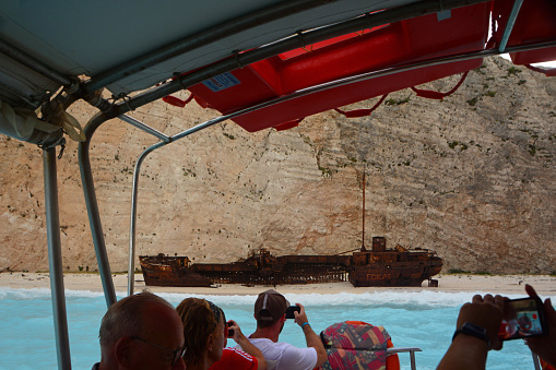 zakynthos-greece 4 -September  / 2023: Navagio Beach; a Tourboat with passengers in the bay where the famous shipwreck of Zakynthos is located in a bay. Surrounded by rock walls. You look at the boat wreck and parts of the people.