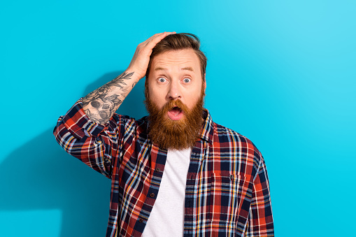 Photo of handsome funny young businessman bearded with tattoo his arm touch head oops mistake confused isolated on blue color background.