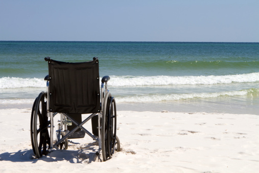Empty wheelchair sits vacant on a beach of sand with ocean waves and surf in the background.
