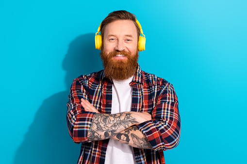 Photo of attractive millennial man red hair beard handsome model listening wireless earphones folded arms isolated on blue color background.