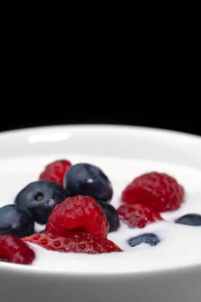 Photo of Close-up of a bowl with natural white yogurt and fresh raspberries, blueberries, strawberries on a black background, macro photography. Delicious healthy breakfast. Vertical photo