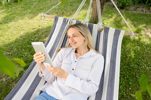 beautiful young woman lying in hammock and working with her tablet, in garden, park