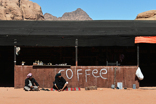 Two Bedouin men sit outside a coffee shop in the desert in Wadi Rum, offering coffee and souvenirs to visitors.