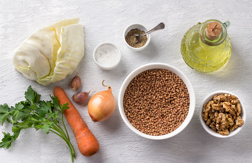 Ingredients for cooking vegan buckwheat with vegetables: buckwheat, cabbage, carrots, onions, garlic, walnuts, parsley, salt, pepper, vegetable oil on a light gray background, top view
