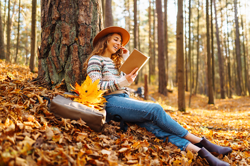 A tourist female in a hat sits in the autumn forest on yellow leaves and reads a book. Beautiful woman enjoying sunny autumn weather.