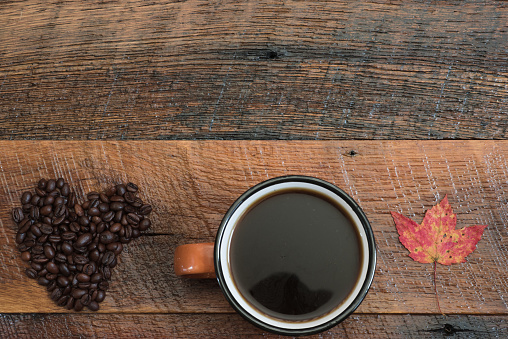 Coffee beans shaped into a heart shape, cup of hot coffee, and a colorful autumn leaf on a rustic wooden table