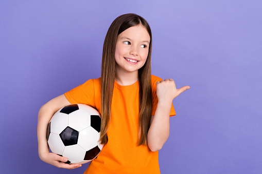Photo of adorable schoolkid dressed orange t-shirt hold soccer ball directing look empty space isolated on purple color background.