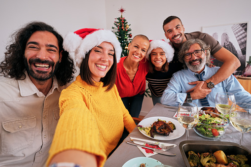 Mobile selfie of smiling Caucasian family looking at camera and happy celebrating Christmas holidays at festive table. Cheerful people posing for photo eating together at home on Thanksgiving Day.