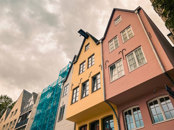 Looking up at multi-colored European style architecture in the city square of Cologne, Germany. One building is covered in scaffolding. Peaked roofs reach towards a cloudy, overcast sky.