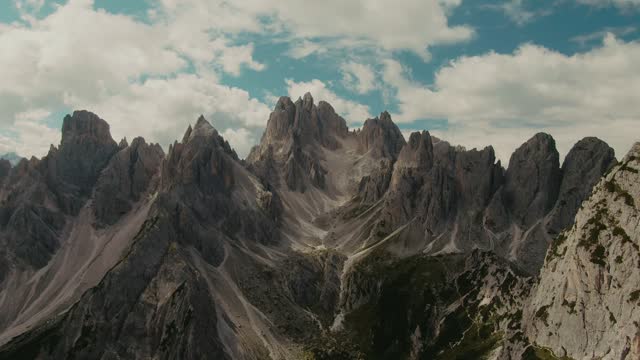 Drone shot with tall steep rocky mountains and partly clouded sky in the background, hiking in the Alps, majestic landscape,  cinematic color grade