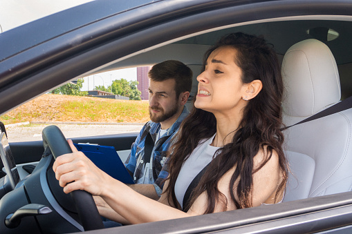 Stressed young woman taking a driving test, male instructor sitting nearby in the car. Test drive, transportation, safety, education concept