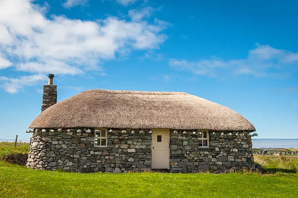 Traditional stone crofter's cottage with stones weighing down the thatched roof in a picturesque summer landscape of green pasture and rolling hills overlooking the mountains of the Western Isles deep in the Highlands of Scotland, UK. ProPhoto RGB profile for maximum color fidelity and gamut.