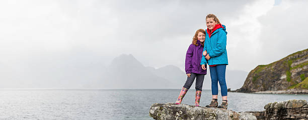 Children in bright outdoor clothing beside ocean Two young girls standing on a rock smiling at the camera overlooking a misty ocean island shore dressed in colourful outdoor clothing on Elgol Beach, Isle of Skye in the Highlands of Scotland. ProPhoto RGB profile for maximum color fidelity and gamut. raincoat photos stock pictures, royalty-free photos & images