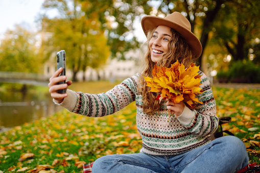 A cheerful woman in a hat sits on a blanket in an autumn park with a phone in her hands, takes a selfie, talks on a video call, blogs. Concept of enjoying nature.