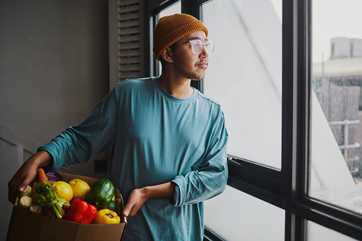 Image of a young man standing by a window, holding a box of freshly delivered fruit and vegetable. Stock photo