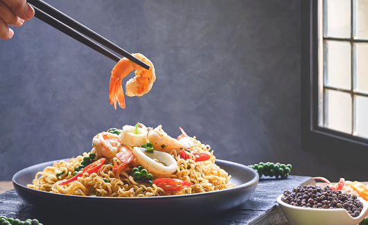 Hand using chopstick picking up shrimp with stir fried instant noodles spicy seafood in black plate in vintage kitchen room, close up with copy space