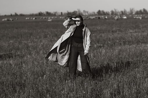 Stylish woman in a coat on a field among dry grass, enjoying nature. Black and white photo. Concept of nature, fashion, freedom. Lifestyle.
