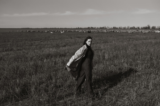 Stylish woman in a coat on a field among dry grass, enjoying nature. Black and white photo. Concept of nature, fashion, freedom. Lifestyle.