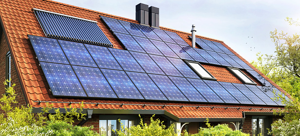 House in residential area has a roof with solar power panels to help generate energy for the family living there. The house has a lawn and a driveway. Concept of using sustainable ressources in everyday life to generate electricity.