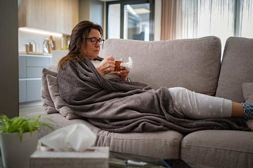 Healthcare and medicine. Woman resting on sofa at home drinking hot tea. She is wrapped in blanket. High resolution 42Mp indoors digital capture taken with SONY A7rII and Zeiss Batis 40mm F2.0 CF lens
