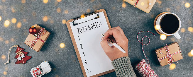 Happy New Year 2024. Woman's hand writing 2024 Goals in notebook. Laptop, present box, cup of coffee, holiday decoration. Top view, flat lay. Banner image for web site.