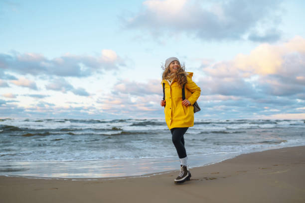 A female traveler in a yellow coat and backpack walks and has fun on the ocean shore, enjoying the scenery. The concept of freedom, travel, recreation. Lifestyle.