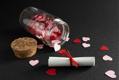 A small paper scroll tied with a red thread. In the background is a small glass bottle with confetti hearts.