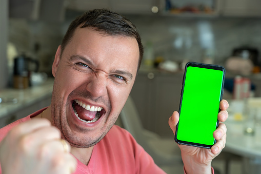 Excited man sits on sofa in living room at home, looks at camera and shows smartphone with green screen for new application. Man sits on couch, holds a smartphone with chroma key mock up green screen