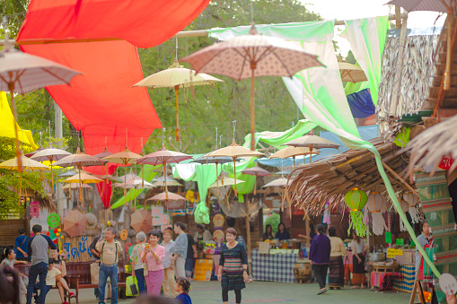 Colorful decorated traditional market in Ratchaburi. Thai people are walking in aisle below umbrella and scarf decor hanging between trees and market stalls.  Market is seated next to temple  โบราณสถานวัดโขลง.  Market offers traditional thai food, thai clothes and other products and music and dance for public and visitors of market.