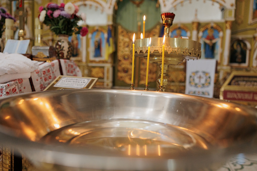 Baptismal font with blessing water and wax candles are against background of Orthodox church interior. Newborns baptism rite in Orthodox church.