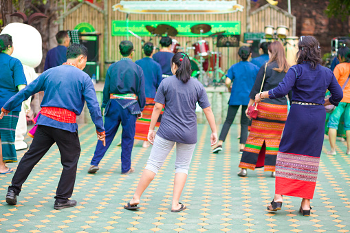 Rear view of dancing thai people captured on traditional market in Ratchaburi. People are wearing traditional clothes. Men are wearing scarfs around hips. Market is seated next to temple  โบราณสถานวัดโขลง.  Market offers traditional thai food, thai clothes and other products and music and dance for public and visitors of market. In background are music instruments of band. One woman in center is trying to dance with people