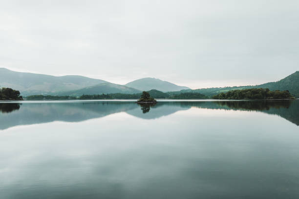 A tranquil view of Kelly Hall Tarn in the Lake District, with the serene pond reflecting the lush meadow and the majestic Old Man of Coniston mountain under a peaceful sky.