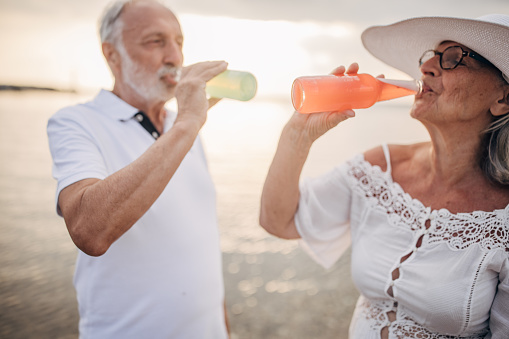 Man and woman, happy senior couple drinking soda on the beach by the sea together.