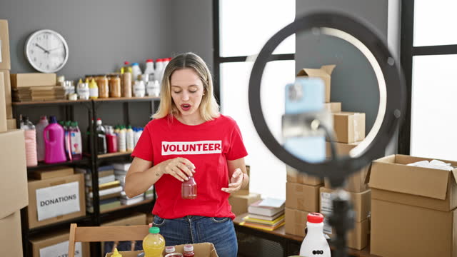Young blonde woman volunteer showing products to donate recording video at charity center