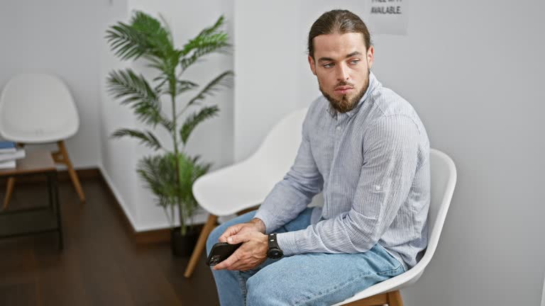 Young hispanic man holding smartphone standing up and walking at waiting room