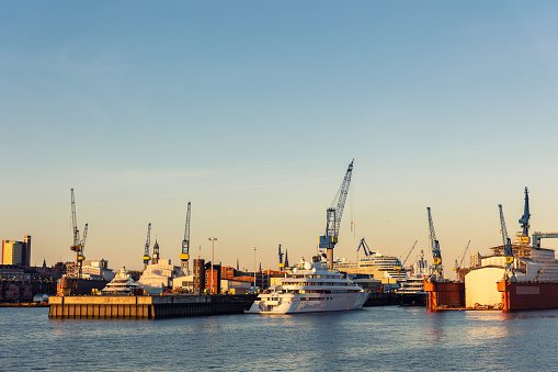 Scenic panoramic view large modern luxury super yacht at maintenance service against construction cargo cranes at dry dock shipyard in Hamburg port sunset sky. Big vessel manufactoring site industry.