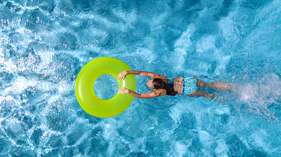 Active young girl in swimming pool aerial top view from above, teenager relaxes and swims on inflatable ring donut and has fun in water on family vacation, tropical holiday resort