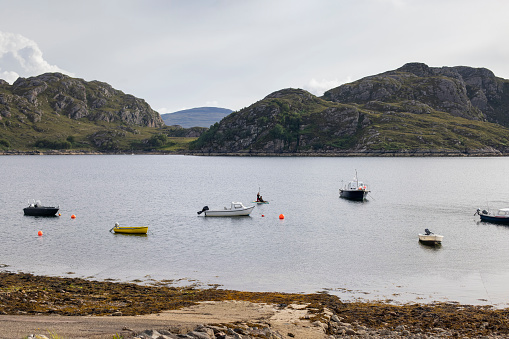A view of the bay at Lower Diabaig in Western Scotland, boats are at anchor, and two girls in wetsuits are playing amongst them, on a paddle board.