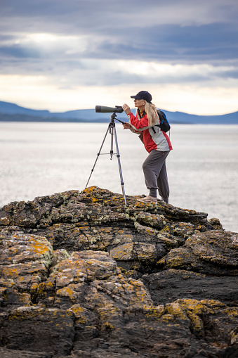 A middle aged female dressed in a red and white waterproof and peaked cap is using a bird watching telescope on a tripod. She is standing on some coastal rocks with the sea (Loch Torridon) and distant hills behind. The area is Lower Diabaig on the western coast of Scotland.